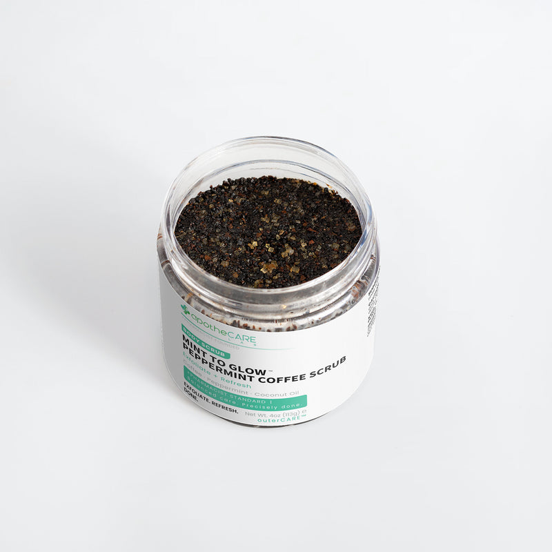 A clear plastic jar filled with dark coffee scrub granules, featuring a white label with green and black text that reads 'Mint to Glow Peppermint Coffee Scrub'.