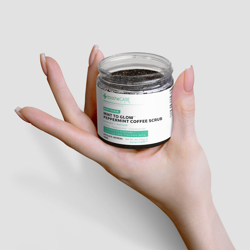 A hand holding a clear jar of Mint to Glow Peppermint Coffee Scrub against a light gray background.