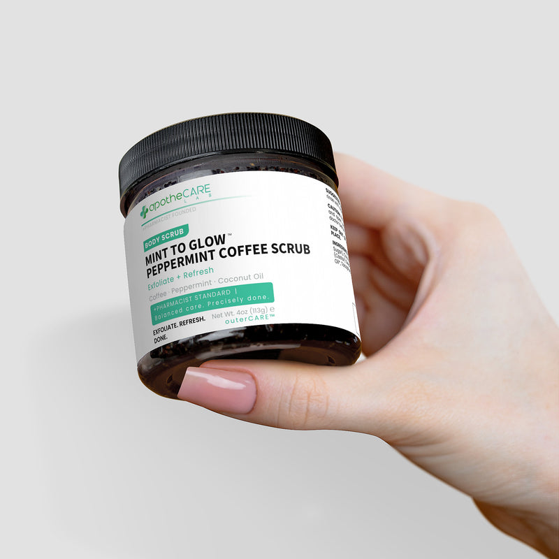 A hand holding a jar of apotheCARE LAB Mint to Glow Peppermint Coffee Scrub against a light gray background.