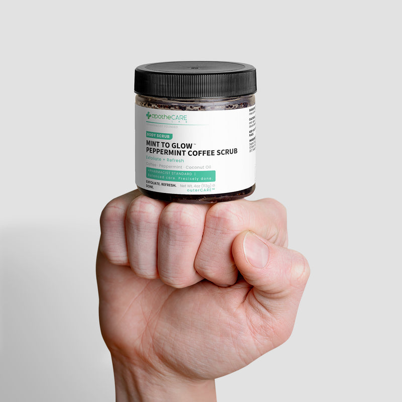 A hand holding a clear jar of Mint to Glow Peppermint Coffee Scrub against a light gray background.