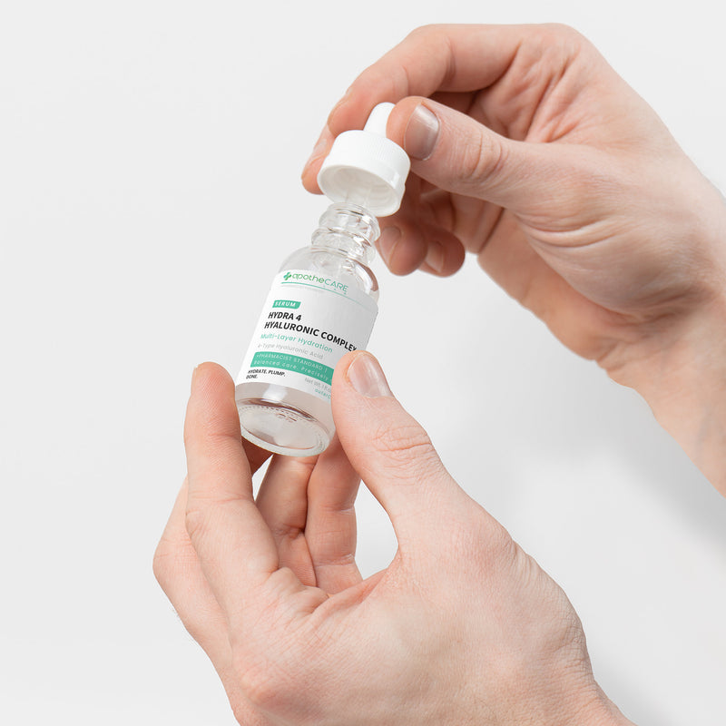 A person's hands holding and unscrewing the cap of a small clear glass dropper bottle of Hyaluronic Serum on a light gray background.