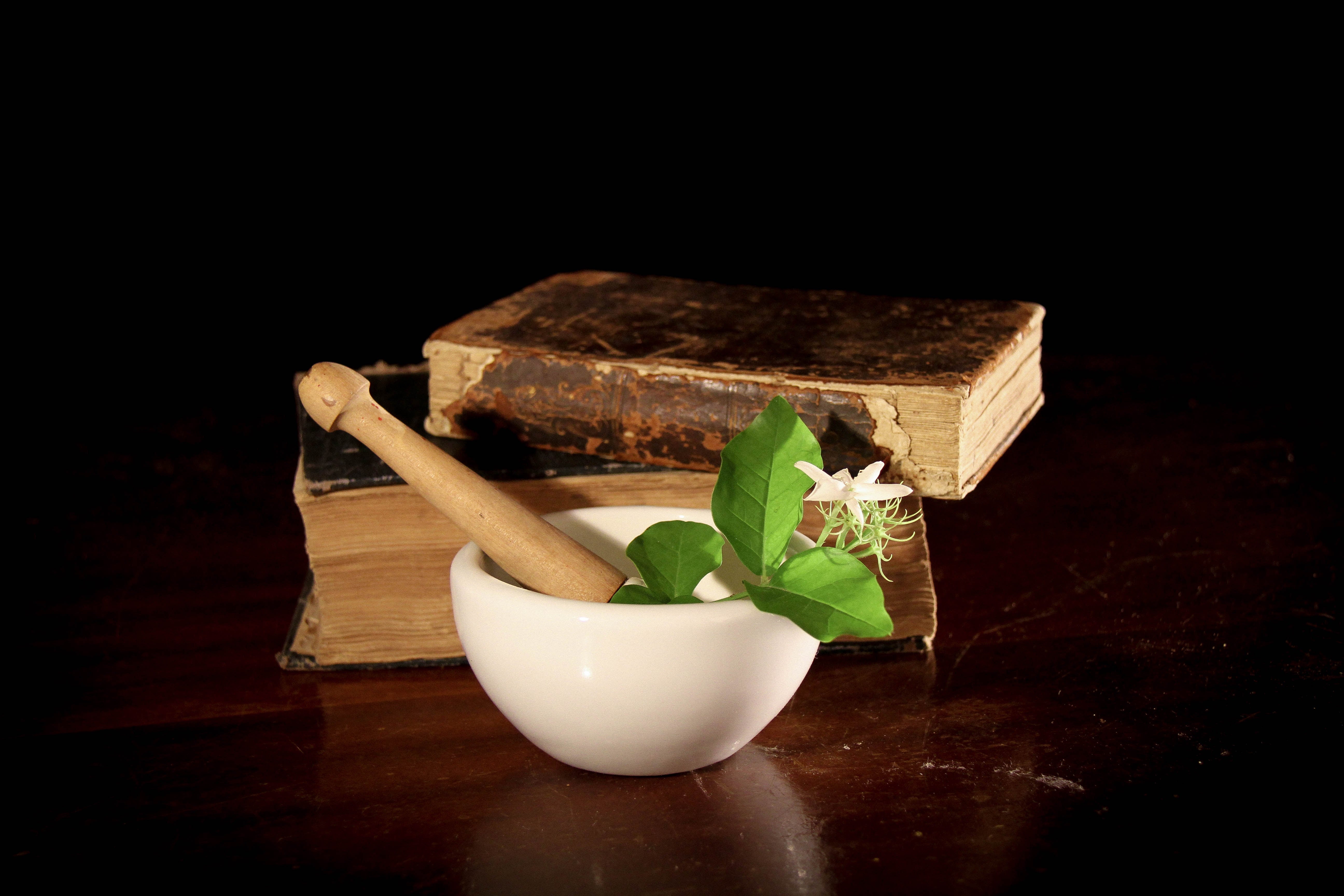 White mortar and pestle with green leaves and flowers on a dark surface with old books.